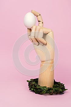 Human wooden arm holding white egg and decorated with spring wreath. Easter composition on pastel pink background