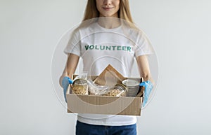 Humanitarian aid concept. Young woman in volunteer t-shirt and medical gloves, holding food donations box, light wall
