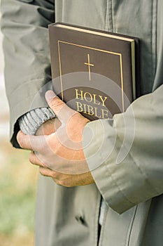 Humble man in light grey coat tightly holding hardcover bible