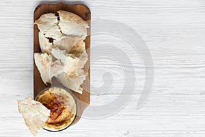 Hummus in bowl with roasted chickpeas, paprika, olive oil and pita bread on rustic wooden board over white wooden background, top