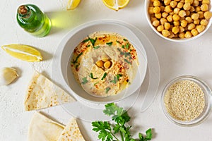 Hummus in a ceramic white bowl with paprika and parsley leaves and ingredients on a light background.