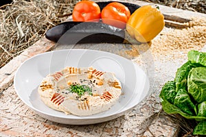 Hummus with greens in a round white plate on a light wooden background among fresh vegetables