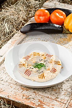 Hummus with greens in a round white plate on a light wooden background among fresh vegetables