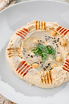 Hummus with greens in a round white plate on a light wooden background among fresh vegetables