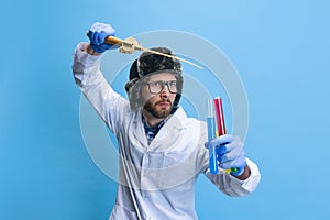 Humorous portrait of young man in image of chemist, doctor wearing white gown and protective gloves isolated on blue
