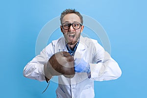 Humorous portrait of young man in image of chemist, doctor wearing white gown and protective gloves isolated on blue