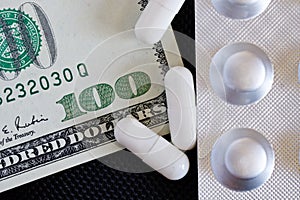 A hundred-dollar bill lies next to white capsule tablets and a silver blister. The problem is the high price of medicines and