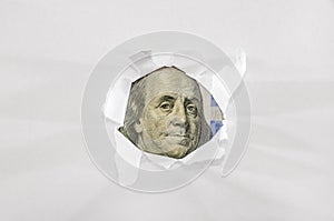 A hundred-dollar bill is visible through a hole in white paper.