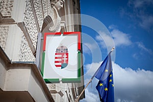 Hungarian coat of arms in front of a flag of European Union waiving in the Hungarian city of Pecs. Hungary is a member of the EU