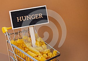 HUNGER text on Blackboard label against Shopping trolley cart Filled With Pasta on Beige background. Food and groceries