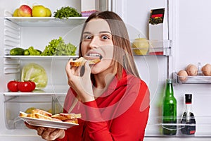 Hungry adorable woman eats delicious sandwhiches with cheese and sausage, stands near opened refrigerator full of products. Beauti