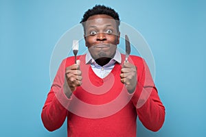 Hungry african man holding fork and knife raised waiting for dinner