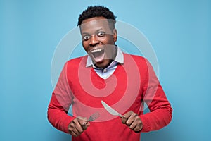 Hungry african man holding fork and knife raised waiting for dinner
