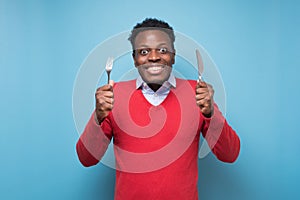 Hungry african young man holding fork and knife on hand ready to eat