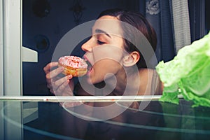 Hungry attractive girl eating donut at night near fridge