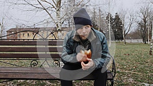 Hungry bearded homeless man sits on a bench and eats a bun in a city park. Below poverty line. Male tramp in dirty