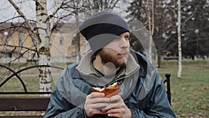 Hungry bearded homeless man sits on a bench and eats a bun in a city park. Below poverty line. Male tramp in dirty