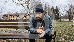 Hungry bearded homeless man sits on a bench and eats a bun in a city park. Below poverty line. Male tramp in dirty clothes with