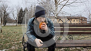 Hungry bearded homeless man sits on a bench and eats a bun in a city park. Below poverty line. Male tramp in dirty clothes with