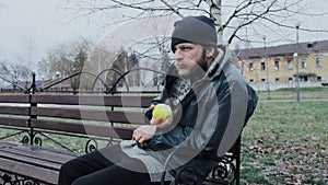 Hungry bearded homeless man sits on a bench and eats a green apple in a city park. Below poverty line. Male tramp in