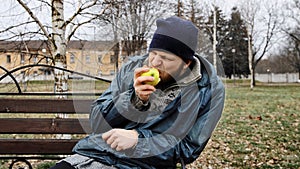 Hungry bearded homeless man sits on a bench and eats a green apple in a city park. Below poverty line. Male tramp in dirty clothes