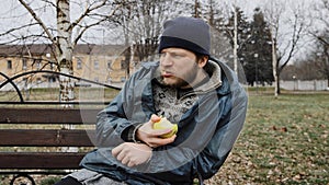 Hungry bearded homeless man sits on a bench and eats a green apple in a city park. Below poverty line. Male tramp in dirty clothes