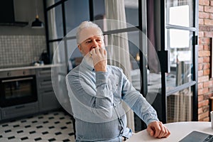 Hungry bearded mature elderly male eating hamburger with beef from fast food restaurant sitting at table and browsing