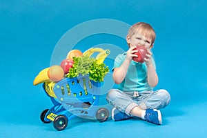 A hungry blond child sits near a supermarket basket full of food carts and bites a huge red apple. The concept of food delivery