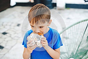 Hungry boy eating a burger at outdoors cafe. Cute child eating fast food