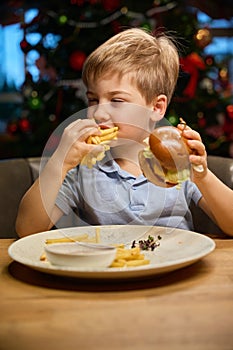 Hungry boy eating delicious burger at festive dinner on Christmas celebration