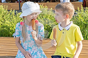 Hungry children eating street food sitting on a park bench. A boy and a girl eating fried sausages on a stick