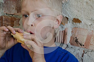 A hungry dirty boy greedily eats a crust of bread against the wall.