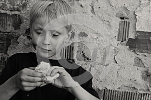 A hungry dirty boy greedily eats a crust of bread against the wall. Black and white.