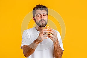 Hungry European Young Man Eating Tasty Burger Against Yellow Backdrop