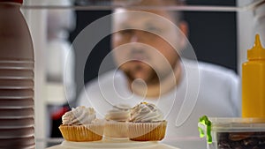 Hungry fat man looking at cream cakes in fridge at night, diabetes risk, sugar