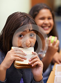 Hungry girl, portrait and student eating sandwich in classroom at school for meal, break or snack time. Young kid or