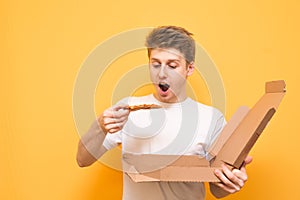 Hungry guy with pizza in his hands is isolated on a yellow background. Emotional young man in a white T-shirt holds in a box and a