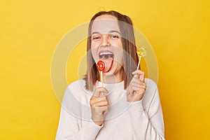 Hungry happy funky teenage girl with ponytails in jumper posing isolated over yellow background licking tasty candy showing tongue