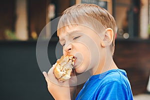 Hungry kid eating a burger at outdoors cafe. Cute child eating fast food. Childhood, unhealthy food concept