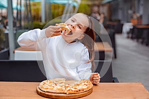 Hungry kid girl in white sweatshirt eating pizza with appetite on cafe terrace. outdoors restaurant copy space