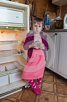 Hungry little girl in old clothes standing next to an empty refrigerator