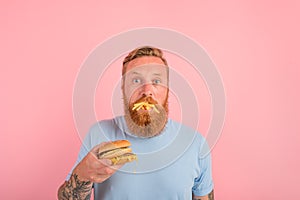 Hungry man with beard and tattoos eats a sandwitch with hamburger and potatoes