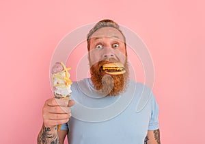 Hungry man with beard and tattoos is undecided if to eat an icecream or a sandwich with hamburger