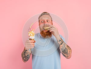 Hungry man with beard and tattoos is undecided if to eat an icecream or a sandwich with hamburger