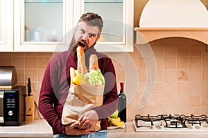 Hungry man bites baguette at kitchen. Bearded man holding paper bag with food at home. Delivery food, products to home