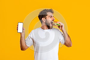 Hungry man with burger shows smartphone blank screen, yellow background