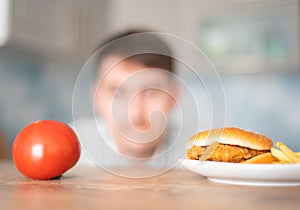 Hungry man chooses between a cheeseburger and a tomato. Fast food or vegetable diet. The temptation of junk food