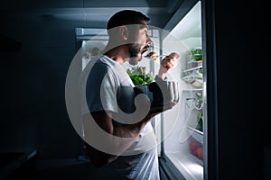 Hungry man eating food at night from open fridge. Man taking midnight snack from refrigerator