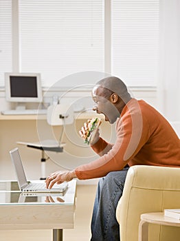 Hungry man eating a sandwich and typing on laptop