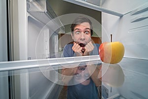 Hungry man is looking for food in fridge and is shocked. Only apple is inside empty fridge